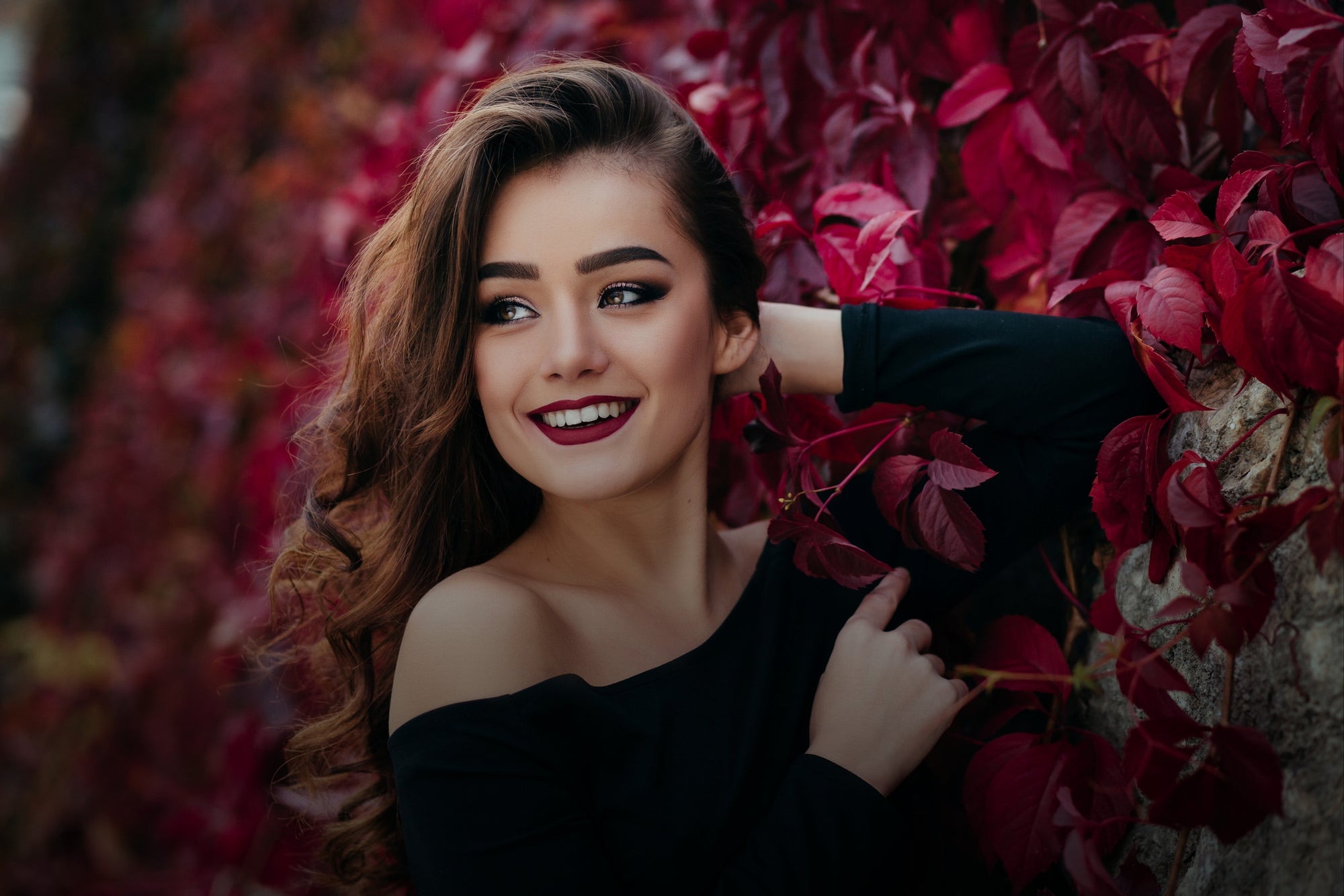 Smiling young woman with dark red lips beside vibrant autumn leaves
