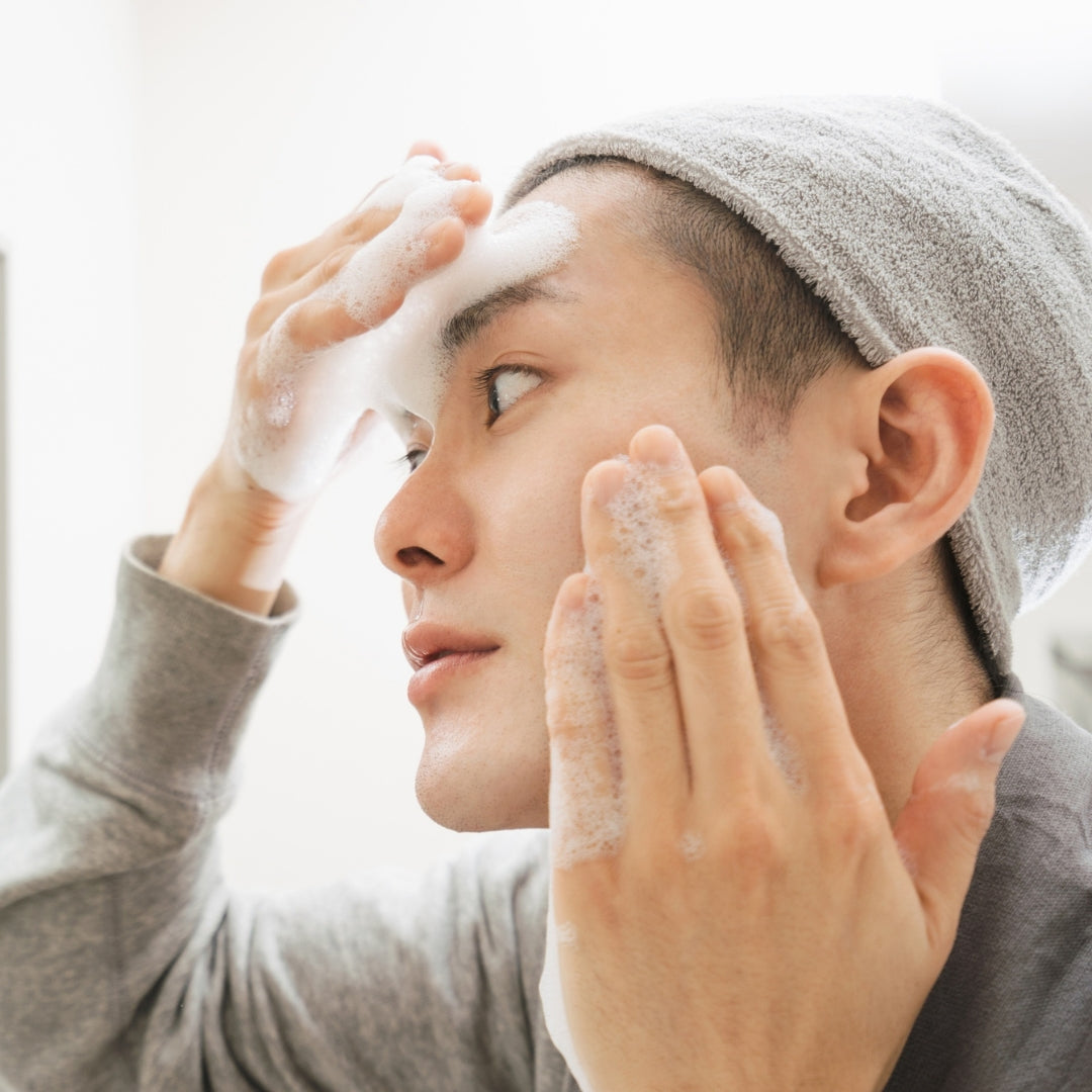 Person washing their face with a towel on their head, wearing a gray sweater.